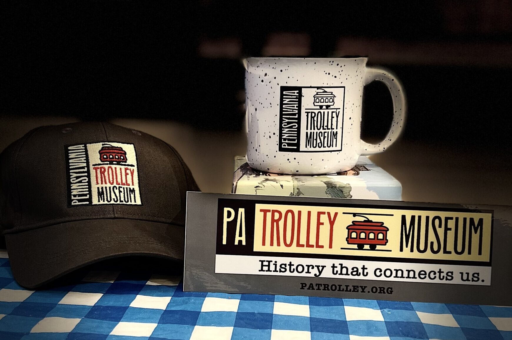 A gray ball cap with the Pennsylvania Trolley Museum logo in the center is at the left of the image, to the front right of the image is a bumper sticker with a gray background and the horizontal version of the Pennsylvania Trolley Museum logo featured. To the back right is a white mug with black speckled paint and the Pennsylvania Trolley Museum logo in all black is featured.