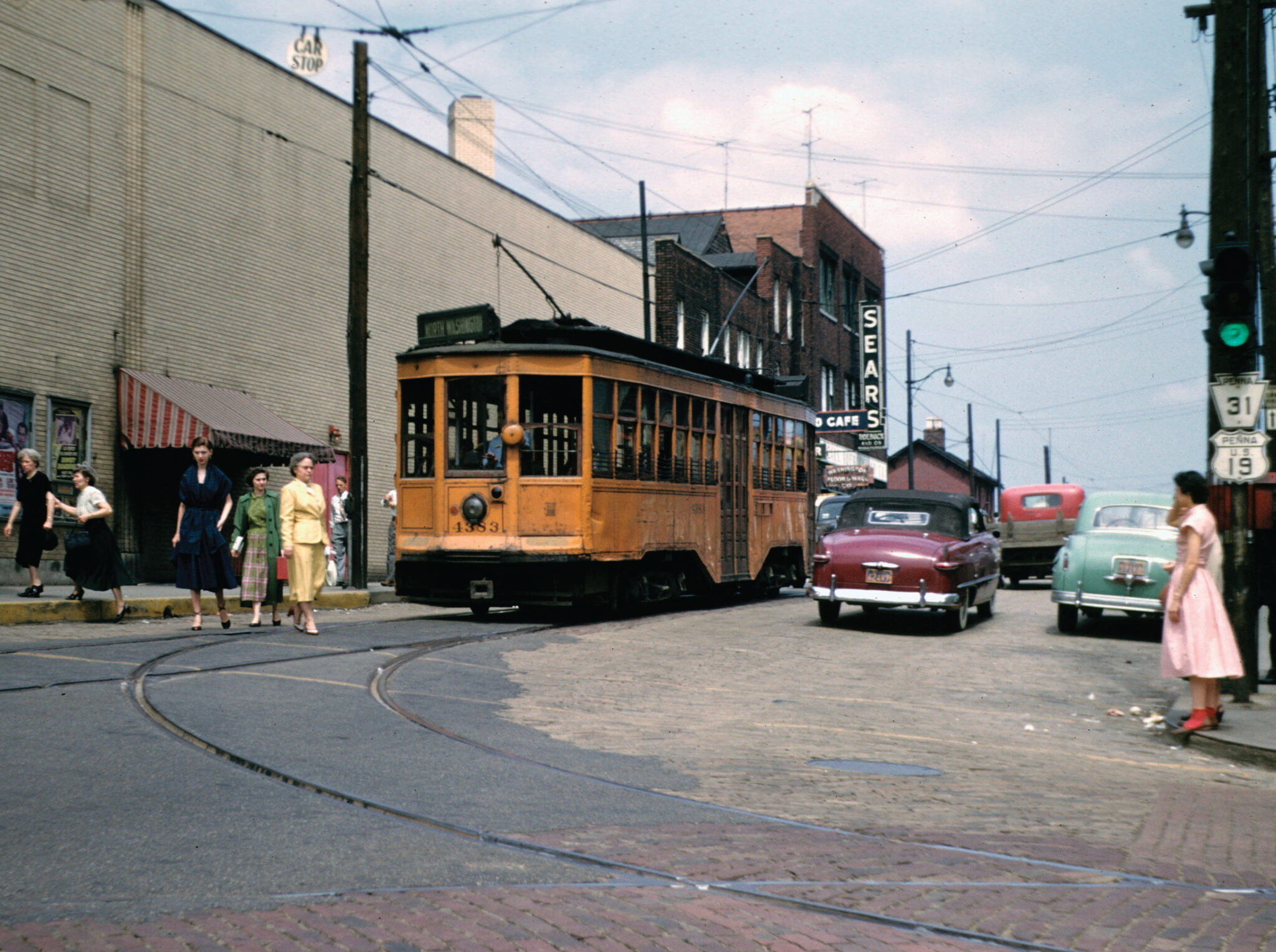 An orange low-floor has just rounded a curve in the rail at the corner of Chestnut St and Main St in Washington, PA in 1952. The trolley is headed away from the photographer. There are people on both sides of the street (behind the trolley), some are beginning to cross the street. There are also cars to the right of the trolley, driving in the street.