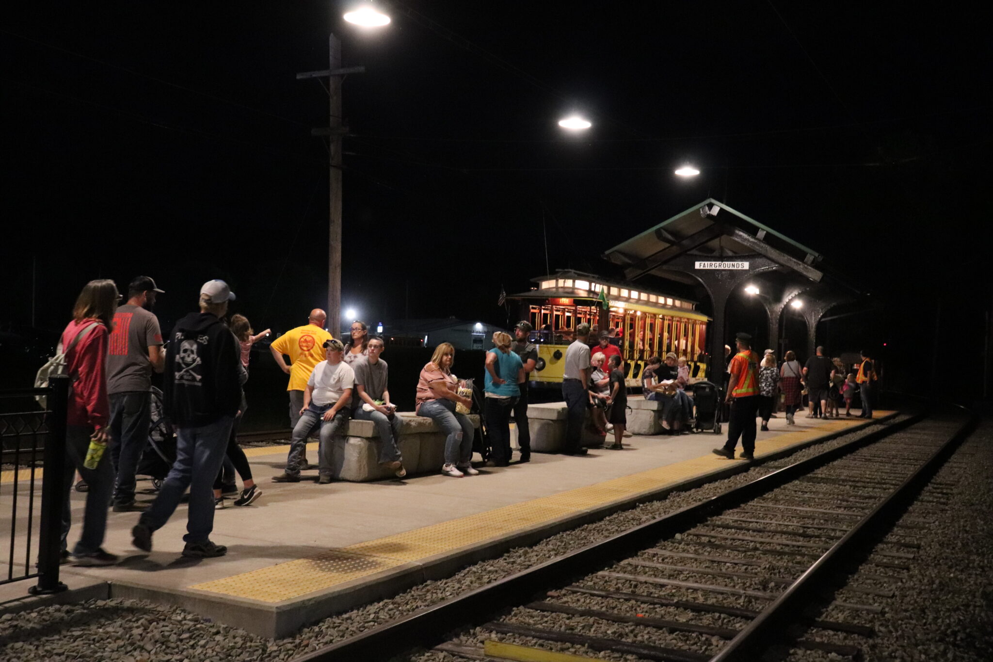 A large group of people wait to board the trolley at the Fairgrounds platform at night. The platform lights illuminate the scene as the open car stops at the far side (in the background of the image).