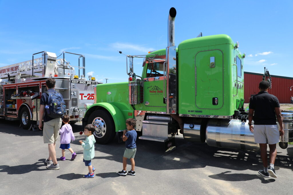 Small children walk with parents alongside a green truck and firetruck